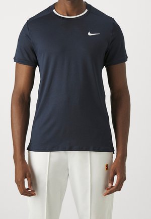 Homme portant un t-shirt Nike bleu marine foncé à manches courtes et un pantalon blanc avec une petite étiquette logo sur la cuisse droite, debout devant un fond uni.