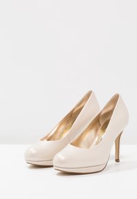 Un paio di décolleté color crema con punta tonda e suola platform, esposte su una superficie bianca con uno sfondo neutro.