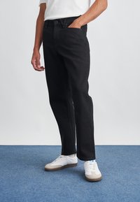 Jeans de mezclilla negra con corte recto, diseño de cinco bolsillos y cierre de botón. Combinados con zapatillas blancas y beige sobre una alfombra azul.