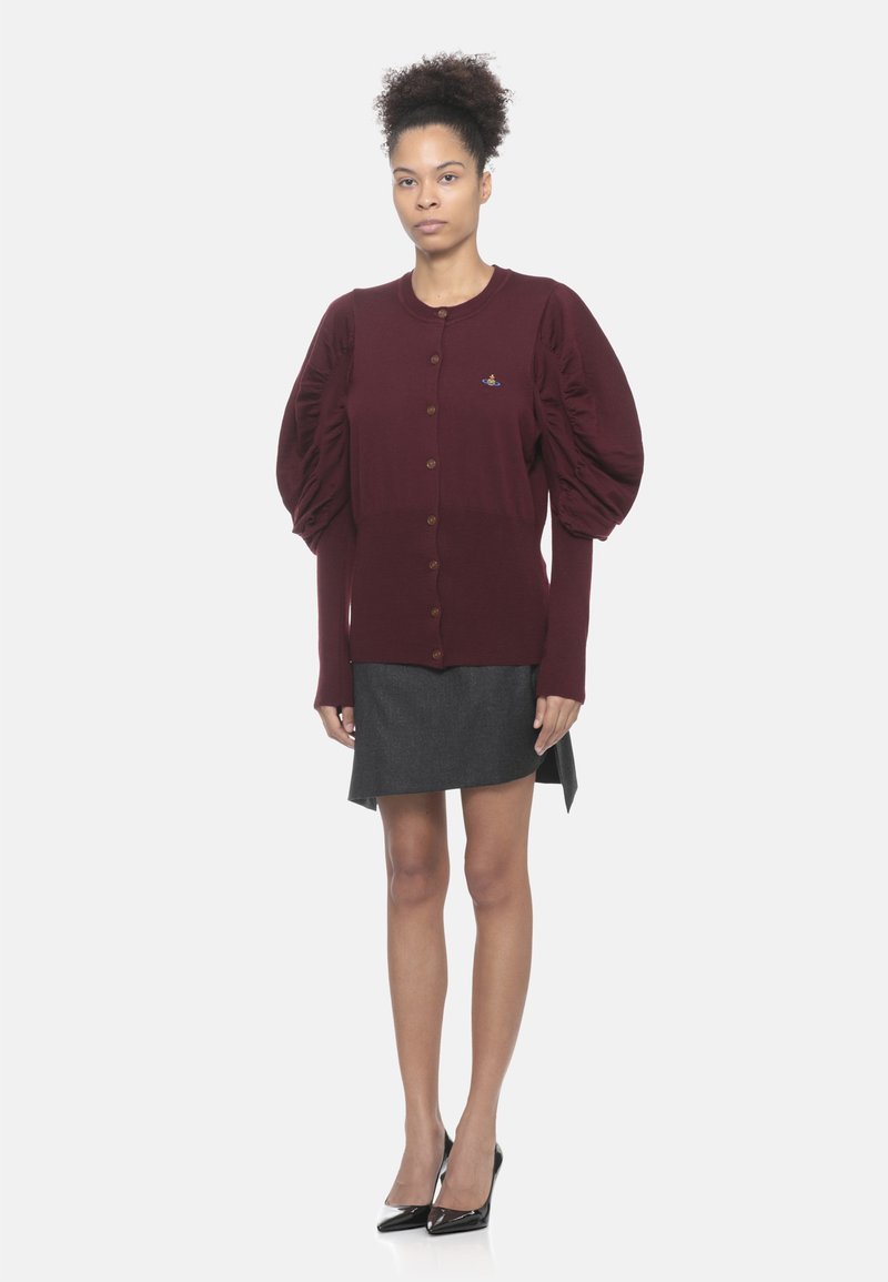 Donna in piedi che indossa un cardigan bordeaux con bottoni e maniche a sbuffo, una gonna grigia scura e tacchi a punta neri su uno sfondo bianco.