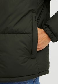 Svart polstret jakke med glidelås-lomme på siden, med en jevn overflate og elastiske mansjetter. Stoffet ser ut til å være vannavstøtende.