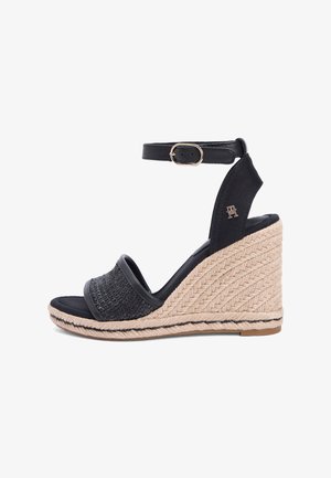 Sandale compensée noire avec bride avant tissée, bride à la cheville avec boucle dorée, et semelle plateforme en style espadrille tressée beige.