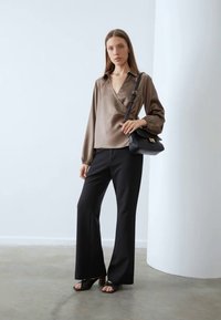 Brown satin wrap blouse, black tailored pants, black shoulder bag, and black heeled mules; model stands against a plain background.