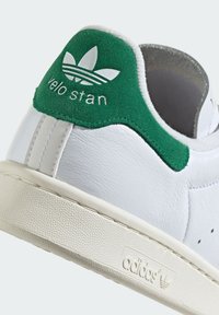 Białe skórzane sneakersy z zielonym zamszowym elementem na pięcie, z napisem "velo stan" oraz trzema białymi paskami. Teksturowana gumowa podeszwa z logo.