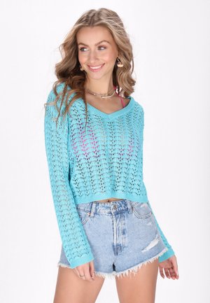 Lyseblå strikket cropped sweater med løs pasform og V-halsdesign, prydet med hulmønstre, parret med slidte denimshorts.