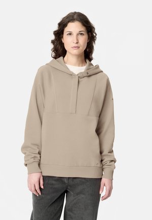 Frau mit lockigem Haar, die einen beigen Hoodie und dunkle Hosen trägt und vor einem schlichten hellen Hintergrund steht.