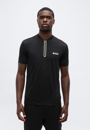 Mężczyzna ubrany w czarną koszulkę z krótkim rękawem i suwakiem oraz logo "BOSS" na piersi, stojący na jednolitym białym tle.