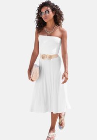 Bijela haljina bez naramenica s lepršavim, dužinom do koljena. Ima dekorativni bež pojas s mašnom, odgovarajuće sandale i pleteni clutch.