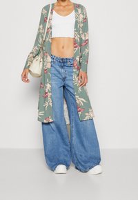 Długi kimono w kwiatowy wzór w kolorze zielonym z różowymi akcentami, noszone na białej bluzce z krótkim rękawem i niebieskich dżinsach z wysokim stanem o szerokich nogawkach. Białe sneakersy.