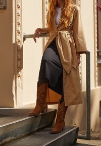 Beżowy trencz do kolan z wiązaniem w talii, czarna spódnica z prążkowaniem oraz brązowe kowbojskie buty z ozdobnym haftem i blokowym obcasem.