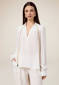 Camicia bianca realizzata in tessuto leggero, con una profonda scollatura a V, maniche lunghe con polsini arricciati e un accento in catena d'oro lungo la parte frontale.