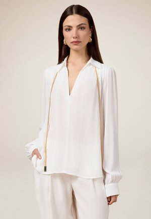 Blusa blanca de tejido ligero, con un escote en V profundo, mangas largas con puños abullonados y un detalle de cadena dorada en la parte frontal.