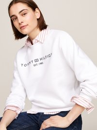 Balta kailinė su mėlynu "TOMMY HILFIGER EST. 1985" logotipu, derinama su raudona ir balta dryžuota marškiniai su apykakle po ja. Džinsai, laisvas kirpimas.