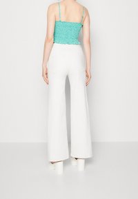 Tyrkysový květinový crop top se spaghetti ramínky a volánkovým lemem, spárovaný s bílými kalhotami širokého střihu. Model má na sobě bílé boty s chunky podpatkem.