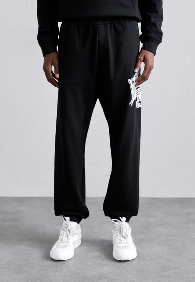 Schwarze Sweatpants mit lockerer Passform und elastischem Bund, die ein weißes grafisches Design am linken Oberschenkel aufweisen. Kombiniert mit weißen Turnschuhen.