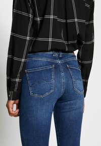 Blå denimjeans med en tight passform, med två bakfickor med sömnadsdetaljer, bärs tillsammans med en svart och vit rutad skjorta.