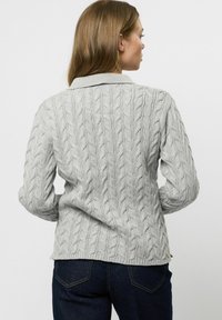 Žena s dlhými hnedými vlasmi, oblečená v svetlosivom svetríku s copánkovým vzorom a tmavomodrých džínsoch, zobrazená zozadu na jednoduchom pozadí.