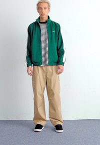 Green jacket with white accents, striped black and white shirt beneath, beige cargo pants, and black sneakers with white soles.