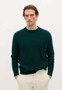 Donkergroen ribgebreid trui met een ronde halslijn en lange mouwen, gecombineerd met lichtbeige broek. De textuur is opvallend en glad.