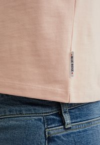 Svetlo roza bombažna majica s kratkimi rokavi in majhno oznako Blue Ridge, z gladko teksturo, v kombinaciji s temno modrimi jeans hlačami.