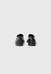 Un par de zapatos de vestir de cuero negro vistos desde atrás, con un fondo gris claro y liso.