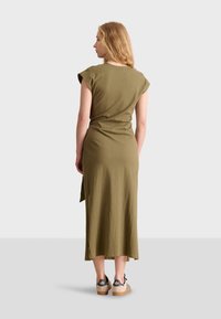 Abito verde oliva, al ginocchio, con maniche a cappuccio, dalla vestibilità leggermente ampia e spacco laterale, abbinato a scarpe con zeppa beige e dettagli neri.