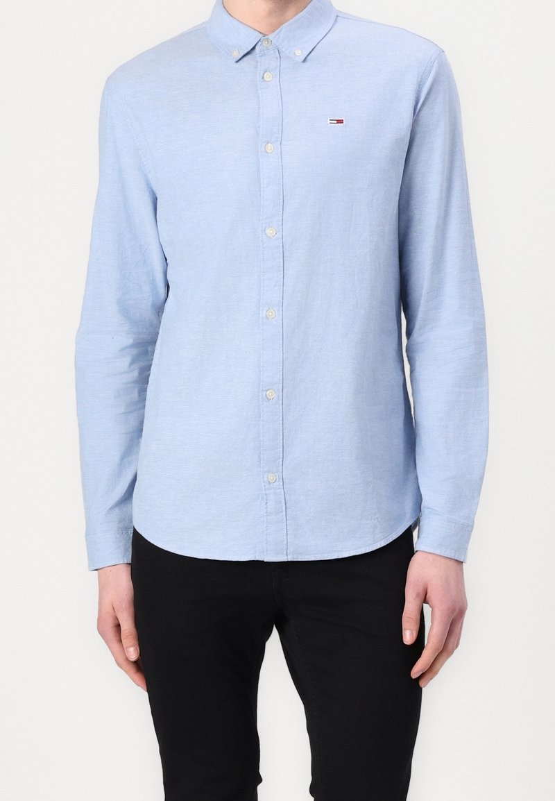 Ljusblå skjorta med knäppning i texturerad tyg, med krage, långa ärmar och en liten logotyp på bröstet. Bärs med svarta byxor.