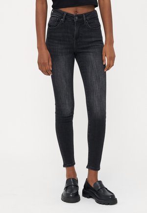 Vrouw draagt zwarte skinny jeans met hoge taille en zwarte leren loafers, staand tegen een effen witte achtergrond.