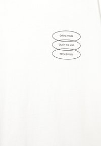 T-shirt bianco in cotone con tre etichette testuali ovali nere: "Modalità offline," "Fuori nella natura," e "Modalità online," disposte verticalmente.
