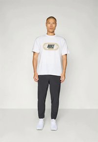 T-shirt branca da Nike com logótipo bege e calças desportivas pretas. O modelo usa ténis brancos. Design desportivo simples e funcional.