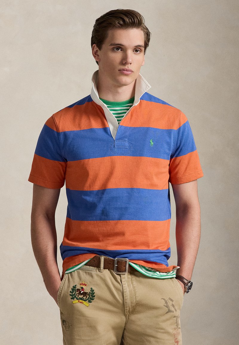 Κοντομάνικο πόλο με οριζόντιες πορτοκαλί και μπλε ρίγες, λευκό γιακά και μικρό πράσινο λογότυπο. Φοριέται με χακί παντελόνι.
