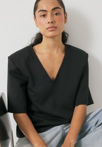 Must mustaga V-kaelusega särk lühikeste varrukatega, valmistatud siledast kangast. Omab struktureeritud disaini ja lamedaid õmblusi. Kandmise ajal koos helesiniste teksadega.