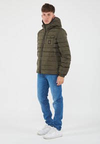 Giacca a puffo verde oliva con chiusura a zip, cappuccio e cuciture orizzontali. Indossata con jeans blu e sneakers bianche.