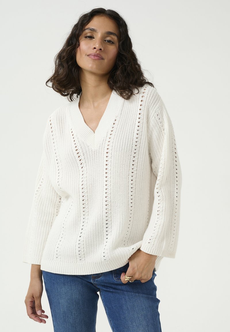Weißer gestrickter Pullover mit V-Ausschnitt, der vertikale Streifen und Lochmuster aufweist. Lockere Passform und heruntergesetzte Schultern. Kombiniert mit blauen Jeans.