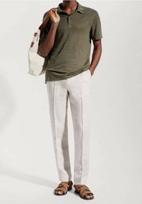 Mann in olivgrünem Polohemd, hellbeigen Hosen und braunen Sandalen, der eine weiße Tragetasche über der Schulter trägt, vor einem schlichten Hintergrund.