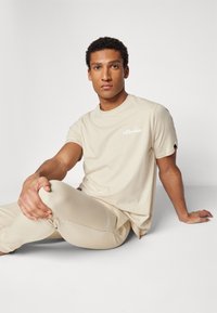 T-shirt en coton beige à manches courtes, arborant un logo blanc sur la poitrine. Le modèle le porte avec un pantalon beige assorti, en position assise.