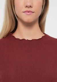 Punainen ribbiknitti toppi, jossa on aaltomainen kaula-aukko ja kimaltava pinta, korostaen tiukkaa muotoilua.