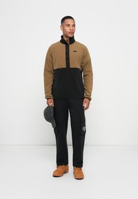 Brun og sort fleece-pullover med halv lynlås og knapfrontdesign. Sorte cargo-bukser og brune støvler, som holder en grå hat.
