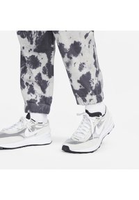 Osoba oblečená v bílých teniskách Nike s černými podrážkami, bílých ponožkách a šedobílých jogger kalhotách s tie-dye vzorem, stojící na bílé ploše.