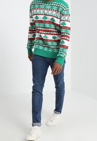 Zielony sweter świąteczny z białymi, czerwonymi i niebieskimi wzorami z motywem choinek i płatków śniegu. Połączony z niebieskimi dżinsami i białymi trampkami.