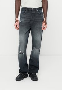 Mørk denimjeans med en falmet finish, som har synlige rifter nær knærne, og en rett bendesign. Brukt med sorte sko.