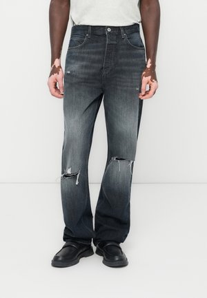 Dark denim jeans with a faded finish, featuring visible rips near the knees, and a straight leg design. Worn with black shoes.
