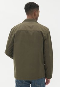 Khaki jakna s ovratnikom in dolgimi rokavi, izdelana iz teksturirane tkanine. Hrbtna stran ima ravno rob in sproščen dizajn, v kombinaciji z modrimi kavbojkami.