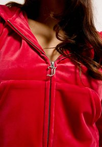 Felpa con zip in velluto rosso, caratterizzata da un tirante argentato con un logo distintivo, progettata con una tasca frontale e un cappuccio accogliente.