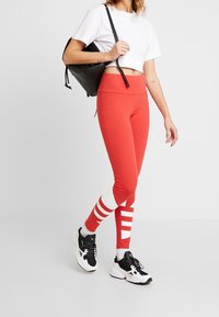 Röda leggings med hög midja, med kontrasterande vita ränder och en mjuk textur, i kombination med en vit kortärmad tröja och en svart ryggsäck.