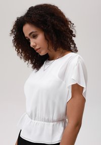 Blusa bianca a maniche corte con vita peplum. Tessuto liscio e semitrasparente, con maniche leggermente arricciate e scollatura rotonda.