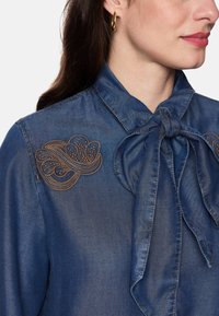 Denim blouse with a bow tie closure, featuring brown embroidered floral accents on the shoulder. Soft texture, deep blue color.