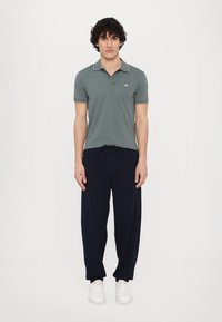 Polo shirt in gedempt groen met contrasterende kraagafwerking, gecombineerd met donkerblauwe trainingsbroeken en witte sneakers. Eenvoudig, ontspannen pasvorm ontwerp.