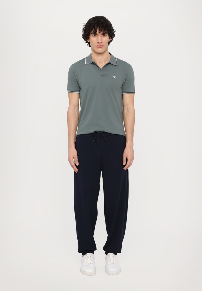 Polo shirt in gedempt groen met contrasterende kraagafwerking, gecombineerd met donkerblauwe trainingsbroeken en witte sneakers. Eenvoudig, ontspannen pasvorm ontwerp.
