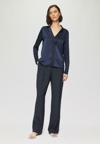 Marine satijnen blouse met lange mouwen en een V-hals, gecombineerd met hoge donkere broeken. Bevat subtiele textuur en een strakke vormgeving.